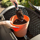 Person adding soil to an orange pot using a garden tool.