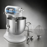 A white stand mixer with a stainless steel bowl is shown on a reflective gray surface against a dark gradient background. The mixer head is slightly open, revealing a glowing blue mechanical detail, with three metal attachments placed beside it: a hook, a whisk, and a paddle.