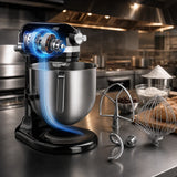 A black and stainless steel stand mixer sits on a commercial kitchen counter with various attachments and baking ingredients nearby, set against a blurred kitchen background.