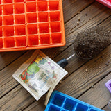 Orange seed tray, soil scoop, seed packet, and blue ice cube tray on a wooden surface.