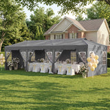 A gray event marquee tent set up on a green lawn in front of a house, decorated with white tablecloths, yellow chair sashes, floral arrangements, and yellow balloons.