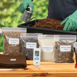 Assortment of vermicomposting materials including bags of worm food and a digital thermometer on a wooden surface.