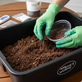 Person wearing green gloves handling earthworms in a container labeled 'Meme's Worms' on a wooden surface.