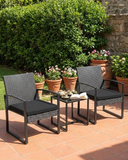Two wicker chairs with black cushions and a small table on a patio with potted plants in the background.