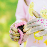 Child's hands wearing gardening gloves holding a pink pot with soil against a blurred green background