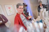 Woman in a red sweater holding a bottle in a room with stacks of boxes and people in the background.
