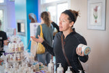 Woman holding a container of ROSEMARY MINT ARNICA SALVE 8 oz. in a store setting with products and people in the background