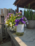 Bottle of lavender essential oil with a purple bow on a wooden surface with flowers in the background.