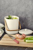 White Astrik Compost Bin on a gray background, surrounded by a cutting board with knife and vegetables.