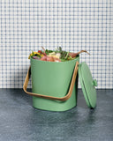 Green Astrik Compost Bin with food waste against a tiled wall background