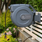 Retractable garden hose reel with nozzle attached, mounted on a wall with greenery in the background.