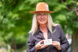 Woman wearing a brown hat and holding a card with a logo, standing outdoors with greenery in the background.