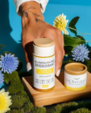 A hand holds a bottle of J+L Naturals Aluminum-Free Deodorant, Vanilla Almond Flavor, next to its open container on a wooden tray, surrounded by blue and yellow flowers against a bright blue background.