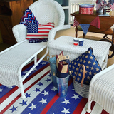 Decorative setting with wicker furniture, American flag-themed cushions, and a table with small cups on a red, white, and blue patterned rug.