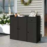 A black wicker-patterned outdoor storage cabinet with two doors and one drawer, shown on a patio in front of a house exterior. The cabinet is styled with small potted plants, books, and a decorative pillow on top.