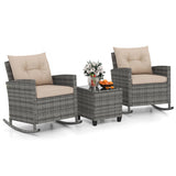 A 3-piece outdoor patio furniture set featuring two gray wicker rocking chairs with beige cushions and a matching gray coffee table with a glass top, styled with a floral arrangement and oranges on a white background.