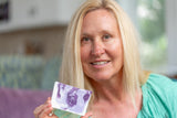 Woman holding LAVENDER GOAT'S MILK SOAP SLICE 5 oz. with a purple design, smiling indoors.