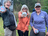 Three women outdoors, holding containers with a logo, in a grassy area.