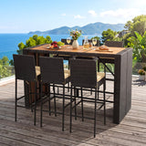 Outdoor high-top dining set on a wooden deck overlooking the ocean, featuring a rectangular table with a wood-look top and dark woven wicker-style sides, surrounded by four matching bar stools with light beige seats.