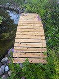 8 ft bridge with no railling extending into a pond with greenery and flowers around