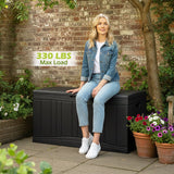 Woman sitting on a large black storage box with a brick wall and plants in the background