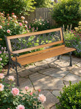A wooden garden bench with a black cast iron decorative backrest and arms, placed on a stone patio in a lush, flower-filled garden.