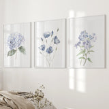 Three floral prints on a wall with a bed and decorative items in the foreground.
