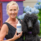 Woman holding a bottle of 'Good Things' product next to a black dog outdoors.