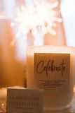 Candle with 'Celebrate' label and 'Homestead Collective' soap in front, blurred lights in the background