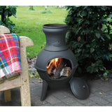 Outdoor chiminea with fire burning next to a wooden bench with a plaid blanket.