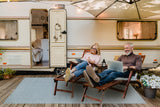 Two people sitting in front of a vintage-style RV, using laptops. Basket Weave Sand & Aqua rug