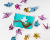 Colorful origami cranes around a BIRD ON BRANCH SOAP SLICE 5.5 oz. on a white background