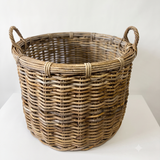 A natural woven wicker basket with two loop handles, shown on a white surface against a white background.