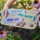 Hand holding a tray with 'Grow Peace', 'Grow Comfort', and 'Grow Love' seed ball packages against a garden background.