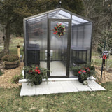 Janco Greenhouse with decorative wreath and potted plants in a garden setting
