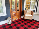 Front porch with a wooden door, wicker chair, and lantern on a red and black Buffalo Plaid Red & Black checkered rug.