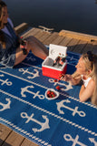 Two women sitting on a blue anchor-patterned rug by a lake, with a cooler and drinks.