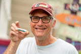 Man wearing a cap and glasses holding a COASTING COCONUT SPF 15 LUBE FOR THE LIPS™ with a blurred background