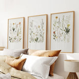 Three framed botanical prints on a wall above a bed with pillows and a lamp.