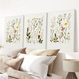 Three botanical wall art pieces above a bed with pillows and a lamp.