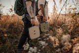Two people walking through a field with backpacks and carrying Nomad Seat | Moss Green