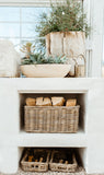 A rustic white stone or concrete fireplace-style shelf with two open cubbies, styled with wicker baskets, stacked firewood, and potted greenery in a bright interior setting.