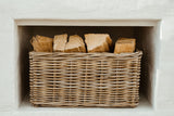 A natural woven wicker basket filled with cut firewood logs, placed inside a white fireplace niche.