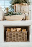 A natural woven wicker basket filled with firewood logs, placed inside a white fireplace or hearth niche. Above the niche, a matching beige bowl planter and rustic wood planter with succulents sit on a white mantel.