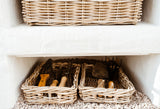 Two natural woven wicker baskets arranged on a white shelf and floor tray, with gardening tools including trowels and a rake resting inside.