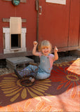 Child sitting on a floral Daisy Rust  rug in front of a red building with a pet door.