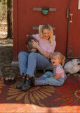Woman and child sitting on a Daisy Rust rug with chickens in front of a red building.