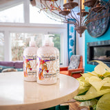 Two bottles of Appalachian Goat Milk Lotion on a table with a decorative chandelier and colorful decor in the background.