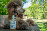 Brown poodle sitting next to a bottle ESB CRAFT BEER SOAP ~ 16 oz. LIQUID SOAP outdoors with trees in the background