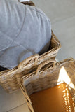A close-up of a woven wicker basket containing a light gray cushion, with a warm light reflecting off the basket's side on a tiled floor.